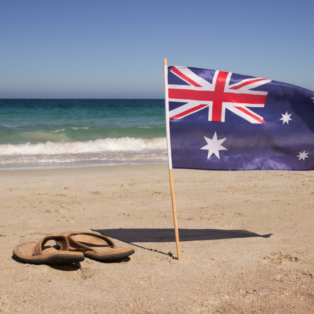 Government Merchandise Supplier Australian Flag at the Beach – Tourism, Community & Council Australian flag in sand on a sunny beach with flip-flops, Australia.