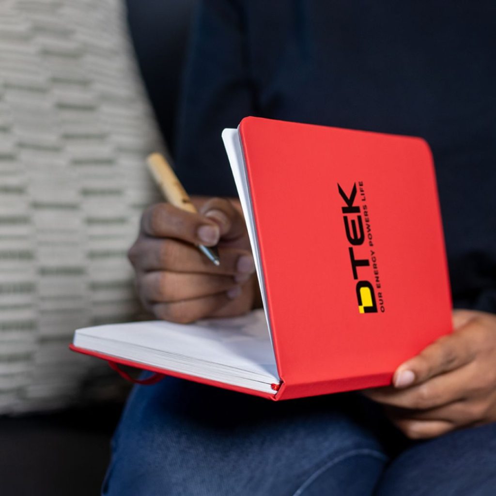 Branded Notebooks for Customer Engagement and Brand Recall Person writing in a red DTEK branded notebook, showing how useful promotional merchandise can keep a brand visible during everyday work.