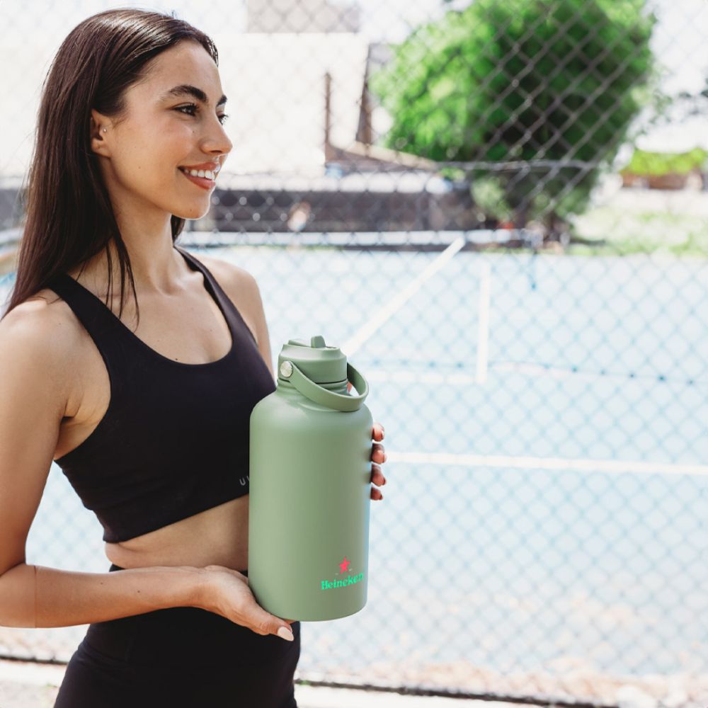 Branded Gym Drink Bottle – Promotional Fitness Merchandise Woman in black activewear holding a large sage green Heineken-branded drink bottle beside an outdoor sports court, showcasing custom promotional fitness merchandise.